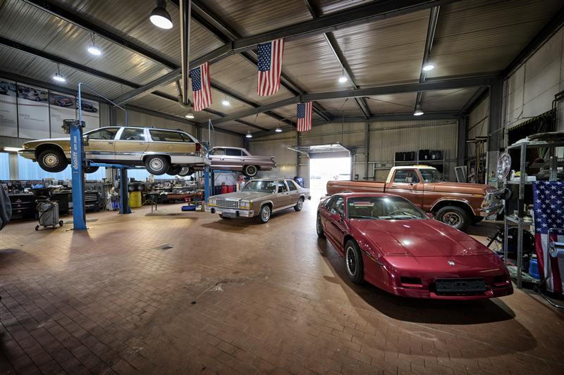Mehrere amerikanische Oldtimer nebeneinander in der Werkstatthalle von Deine Autostube, amerikanische Flaggen unter dem Dach